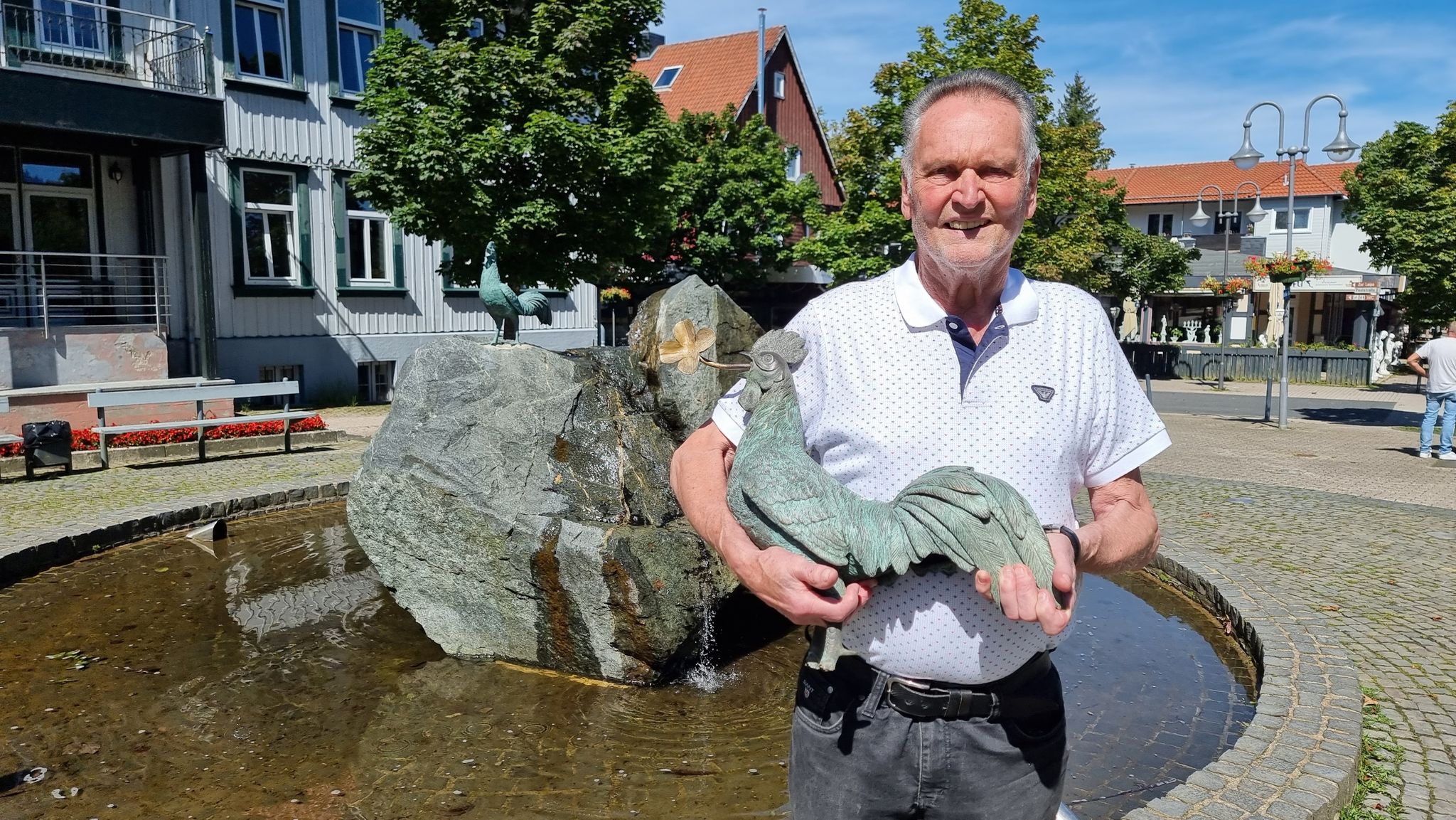 Der Stifter Des Neuen Und Alten Bronze-Hahns, Klaus Elders, Steht Mit Der Figur Vor Einem Brunnen.