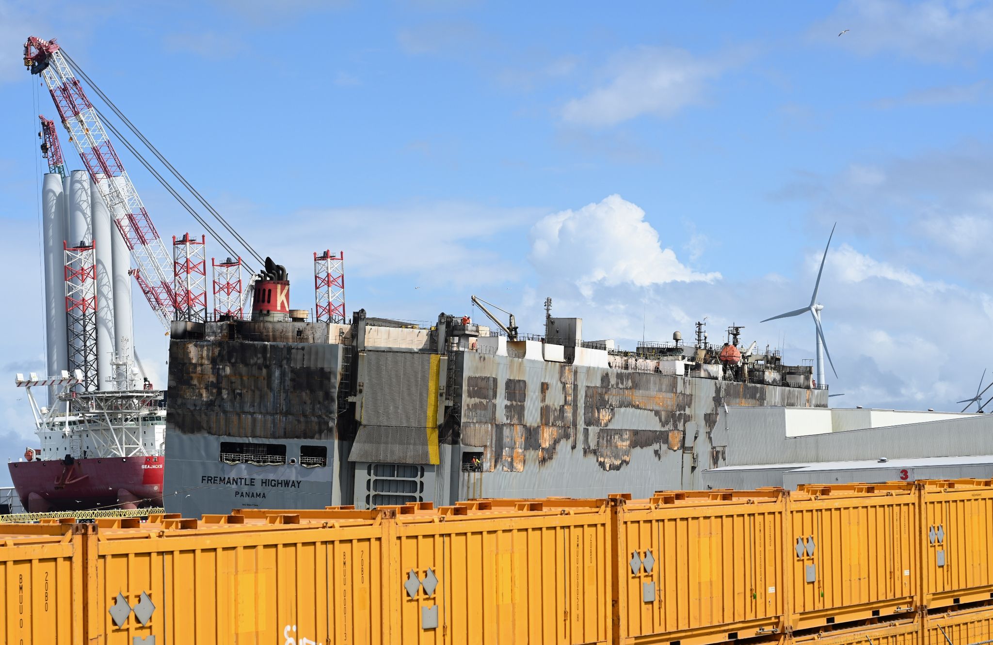 Der Schwer Beschädigte Autofrachter »Fremantle Highway« Liegt Im Hafen, Abgeschirmt Durch Gelbe Container.